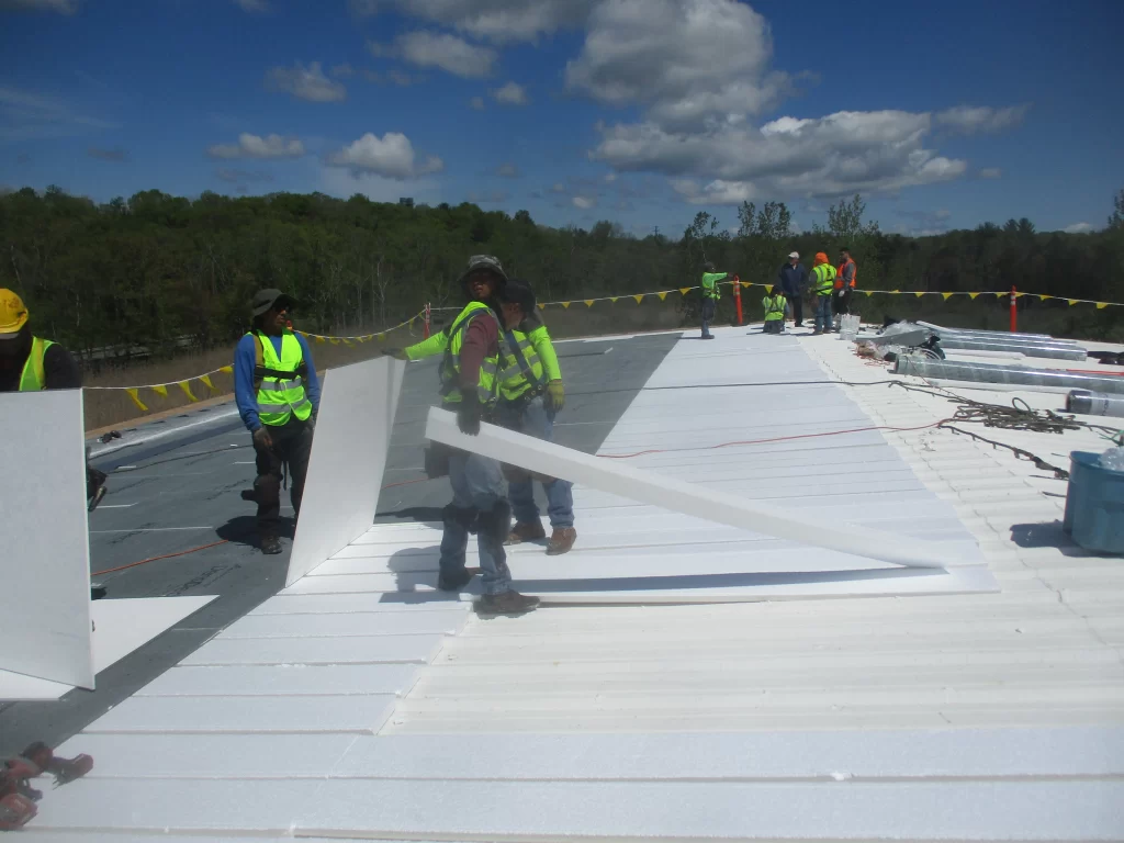 Workers installing white foam insulation boards on commercial flat roof with safety barriers
