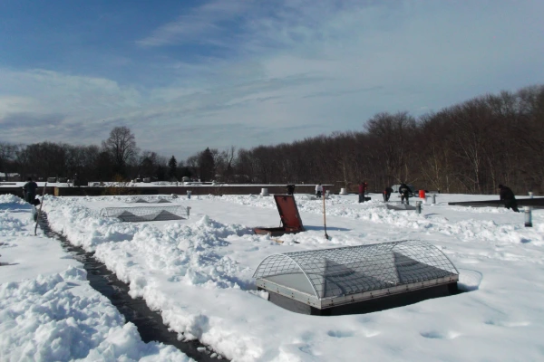 Wide shot of a snow-covered commercial roof with multiple skylights protected by wire cages, and workers performing snow removal in the distance.