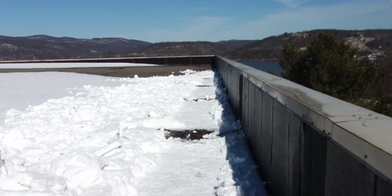 A cleared pathway on a commercial flat roof covered in snow, showing the drain area and a mountainous, water-side landscape in the background.