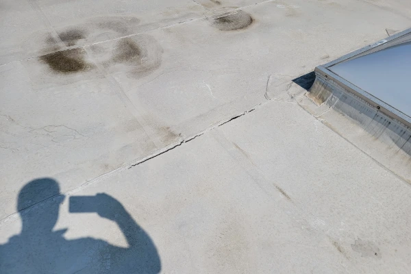 Commercial roof inspector examining a flat roof membrane with visible cracking, dark staining, and damage near a skylight penetration.